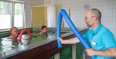 Foto: Zwei Patienten bei der Wassergymnastik, angeleitet durch einen Therapeuten.