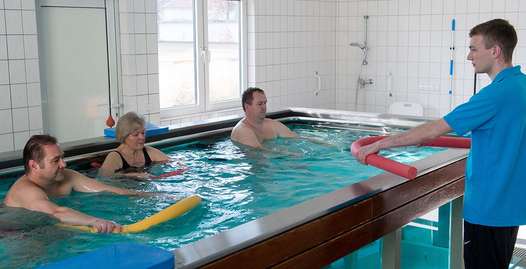 Foto: Drei Patienten bei der Wassergymnastik, angeleitet durch eine Therapeutin.