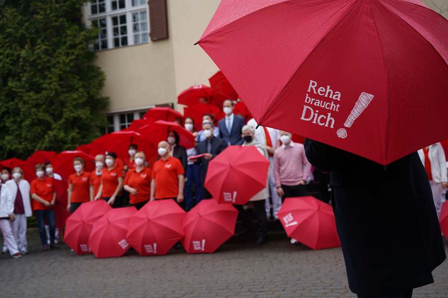 Roter Schirm mit der Aufschrift "Reha braucht Dich" im Vordergrund, dahinter Mitarbeiter der Klinik.