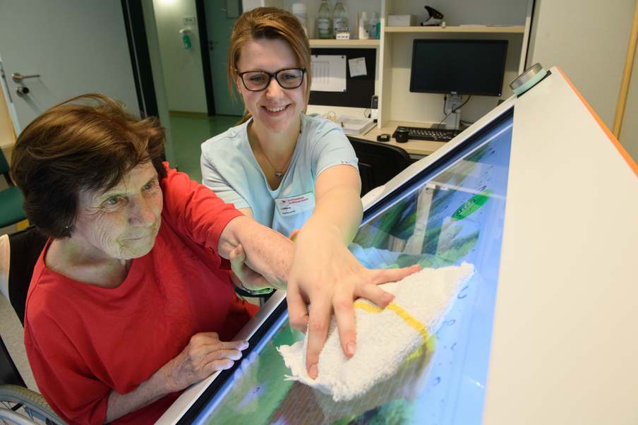 Eine Patientin wird unter Anleitung von einer Therapeutin die Hand geführt bei der Bedienung eines großen Bildschirms.